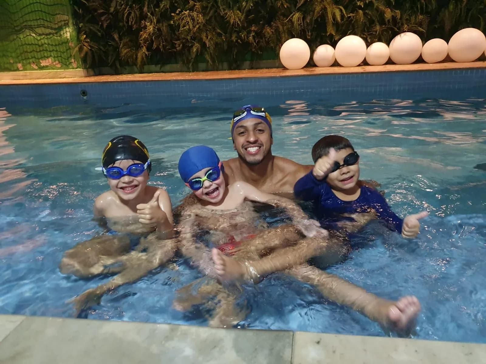 Instructor in the pool with three boys in swim caps and goggles giving a thumbs up