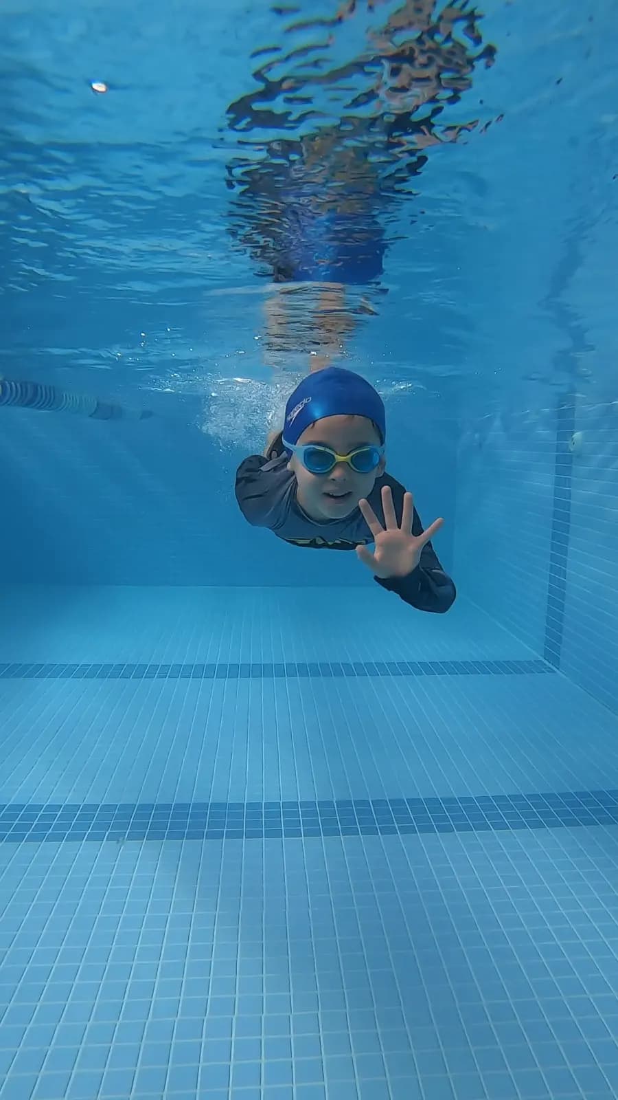 Criança de touca azul e óculos nadando debaixo d’água em direção à câmera