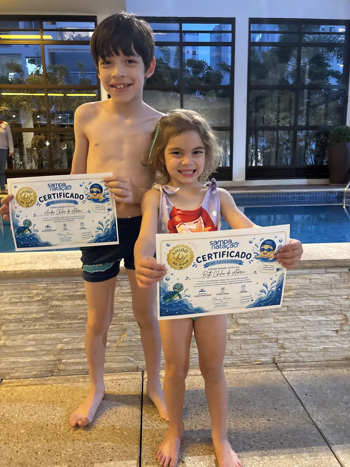 Duas crianças na borda da piscina segurando seus certificados de natação